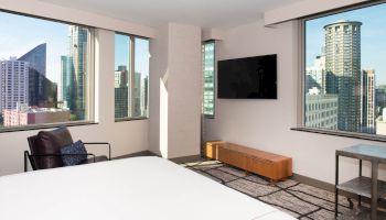 A modern hotel room with large windows displaying a city skyline, featuring a bed, TV, and seating area.