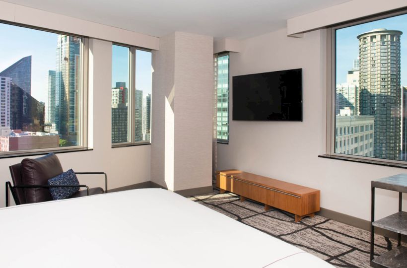 A modern hotel room with large windows displaying a city skyline, featuring a bed, TV, and seating area.