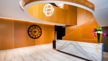 A modern lobby with wooden accents, a large decorative clock, marble reception desk, and hanging spherical lights with flowers.