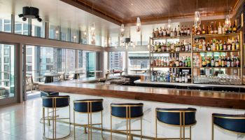 The image shows a modern bar with stools, a wooden counter, and numerous bottles on shelves, situated in a building with large windows.
