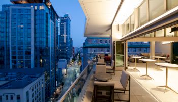 A modern rooftop terrace with seating, overlooking a cityscape of tall buildings at dusk, under a softly lit sky to the left.