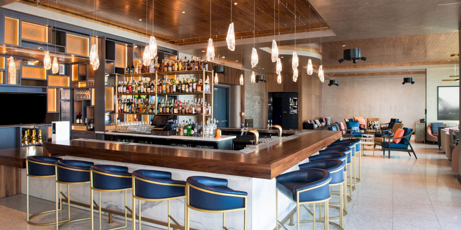 The image shows a modern bar with a wooden counter, blue chairs, hanging lights, and a well-stocked liquor shelf, in a stylish lounge setting.