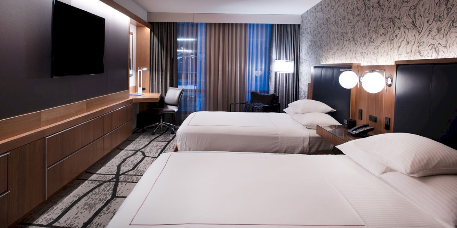 A modern hotel room features two beds, a TV, desk with chair, lamps, and patterned walls and carpet, creating a cozy ambiance.