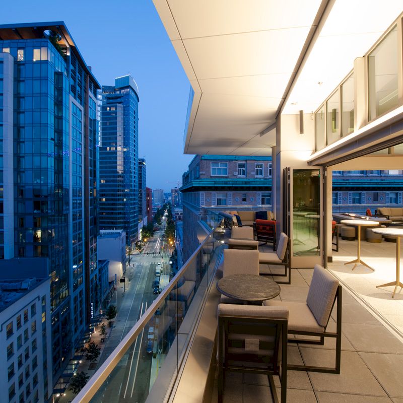 A modern city view from a balcony with tables and chairs, overlooking skyscrapers and a street below at dusk.