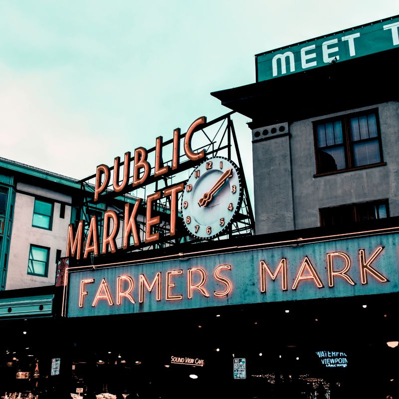 The image shows a public market sign with a clock and a neon "Farmers Market" sign, set against urban buildings under a cloudy sky.