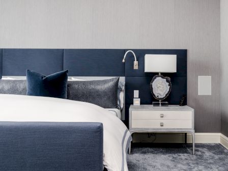A modern bedroom features a navy and white bed, matching pillows, and a nightstand with a lamp and decor on a plush carpet.