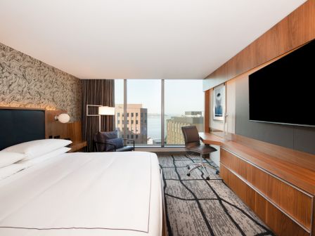A modern hotel room with a large bed, a desk, a chair, and a flat-screen TV. Large windows provide a cityscape view.