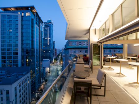 A modern cityscape with tall buildings and a balcony featuring tables and chairs, viewed during the evening.