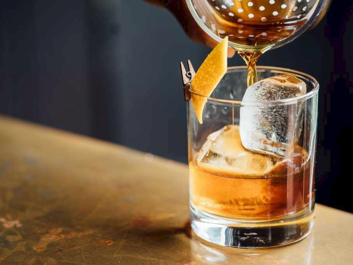 A drink is being poured into a glass with ice and a twist of citrus peel, using a strainer.