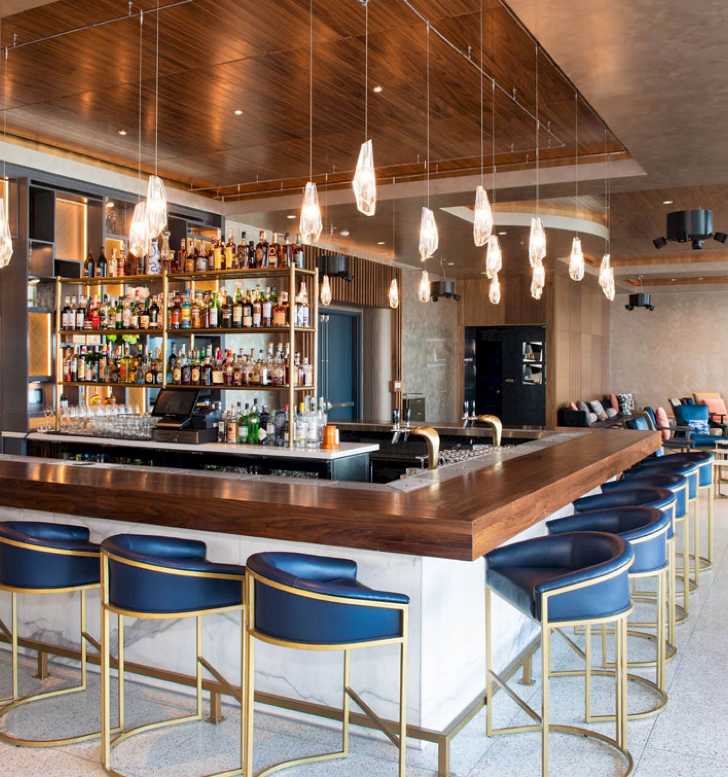 This image shows a modern bar interior with a central counter, stylish seating, pendant lights, and a well-stocked back bar shelf.