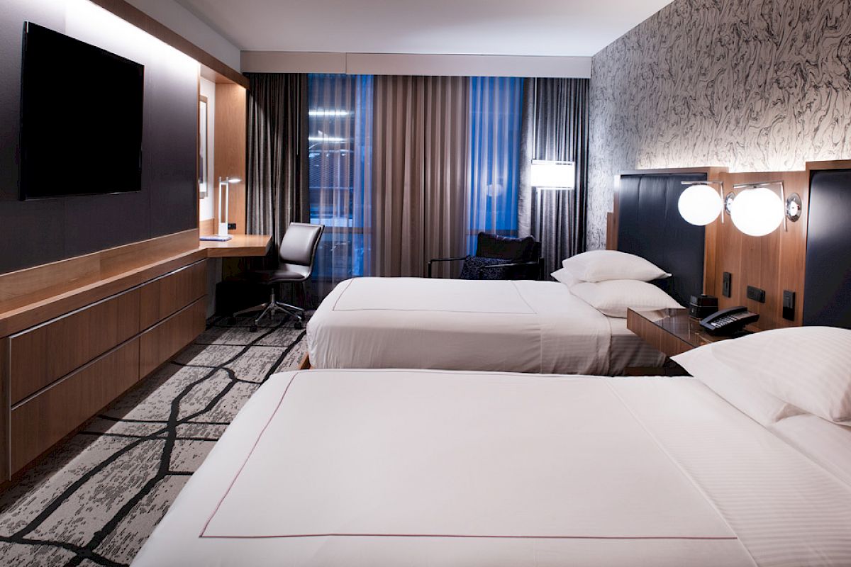 The image shows a modern hotel room with two beds, a TV, a desk, and stylish lighting on a patterned carpet.
