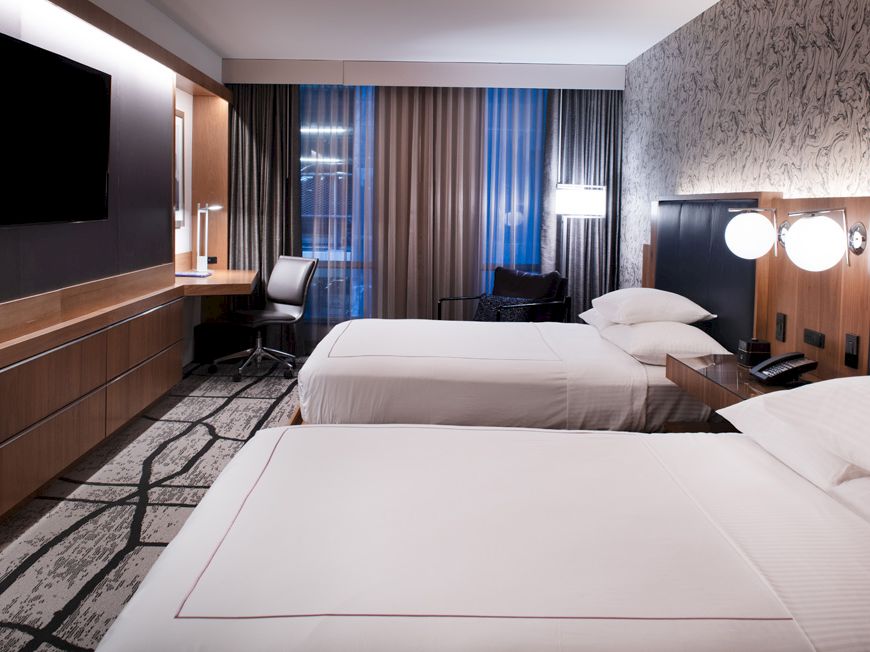 The image shows a modern hotel room with two beds, a TV, a desk, and stylish lighting on a patterned carpet.
