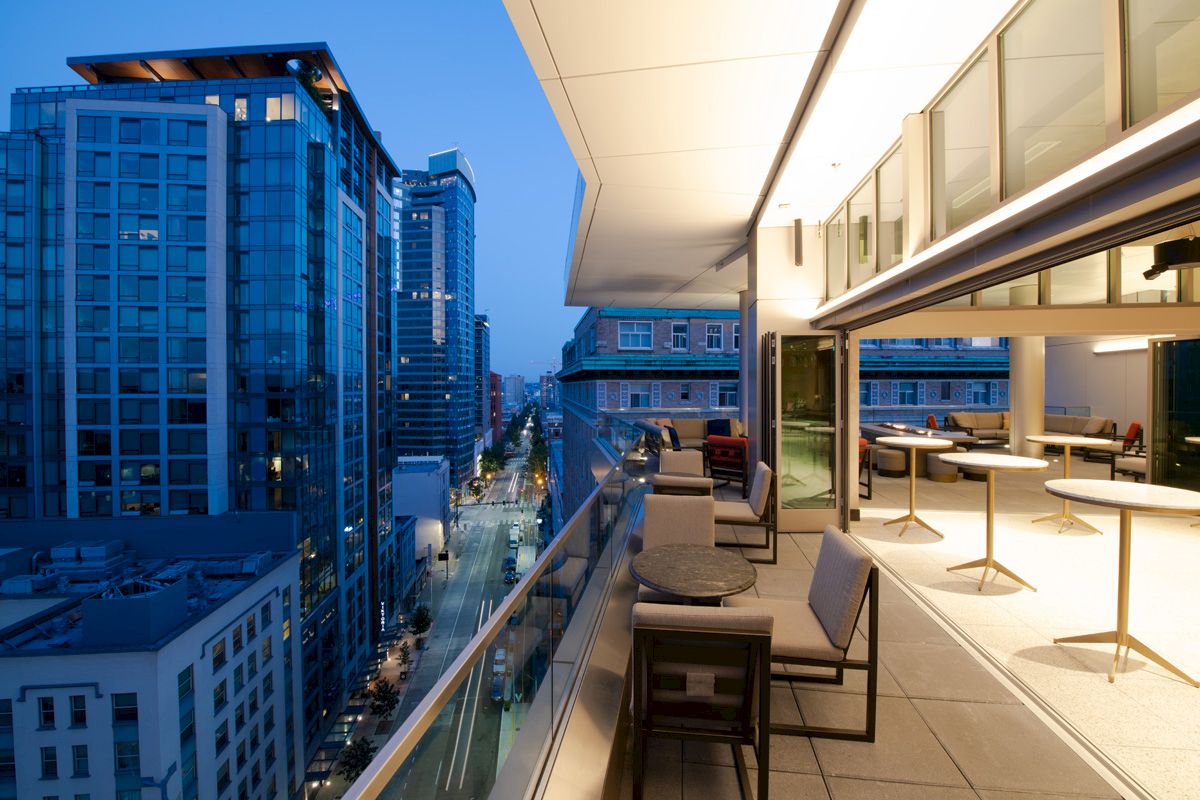 The image shows a modern balcony with seating overlooking an urban cityscape at dusk, featuring tall buildings and city lights.