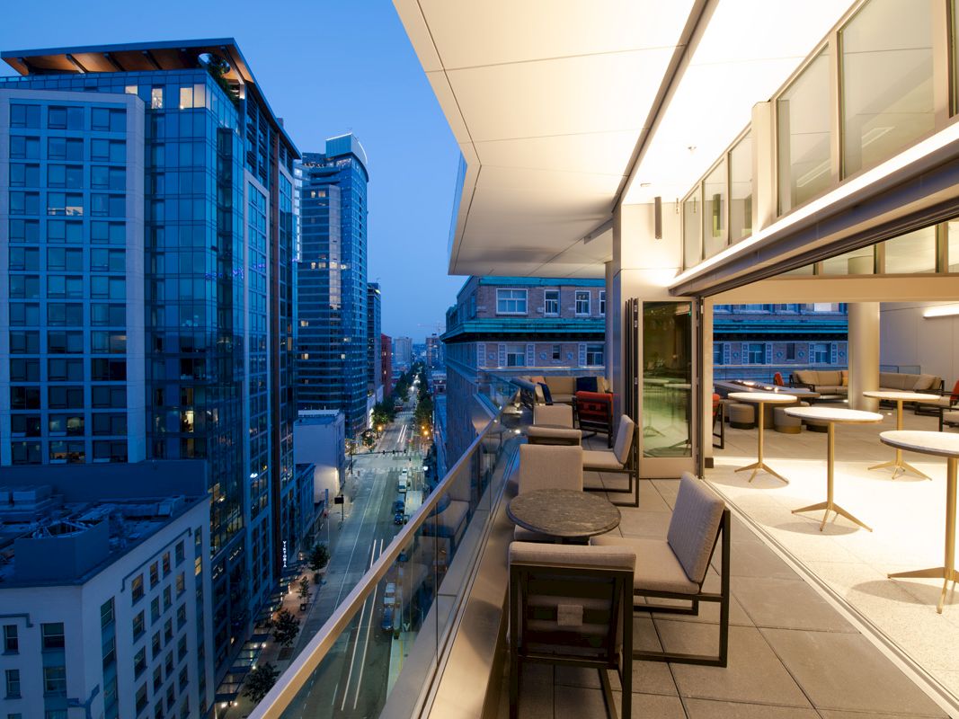 The image shows a modern balcony with seating overlooking an urban cityscape at dusk, featuring tall buildings and city lights.