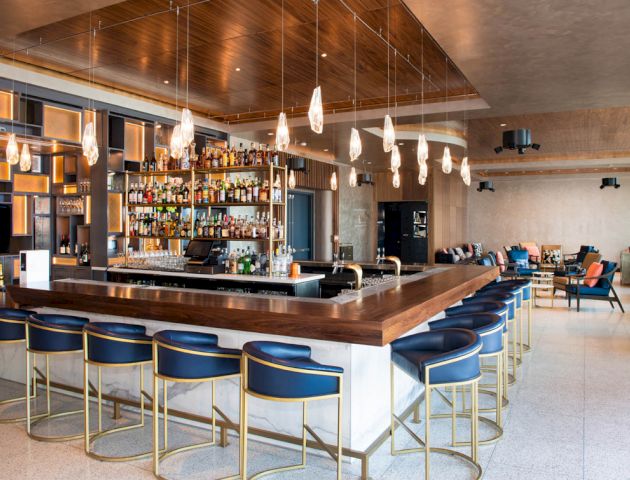 A stylish bar with a wooden counter, blue stools, overhead lighting, and a well-stocked liquor shelf, accompanied by cozy seating.