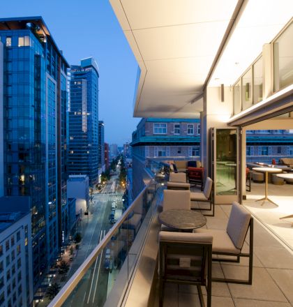 A modern rooftop patio with tables, chairs, and views of a cityscape at dusk. The scene is vibrant with illuminated buildings in the distance.