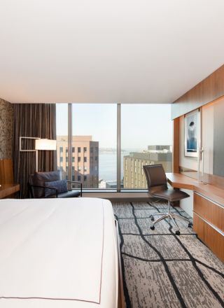 A modern hotel room with a large bed, desk, chair, TV, and window displaying a city view.