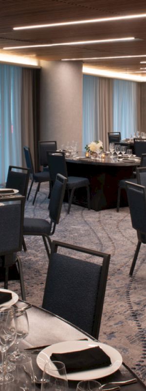 A banquet hall with round tables set for an event, featuring black chairs, white flowers, and glassware on each table.