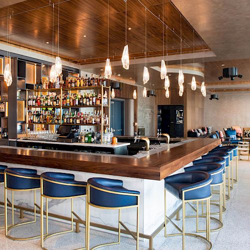 The image shows a modern bar with blue barstools, pendant lights, and a fully stocked liquor shelf, creating an inviting atmosphere.