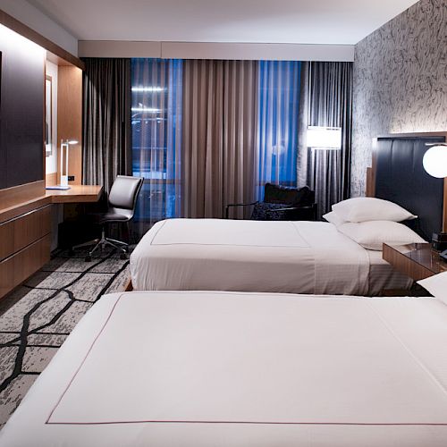 The image shows a modern hotel room with two beds, a flat-screen TV, a desk and chair, lamps, and a stylish patterned carpet and wallpaper.