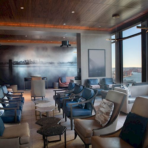 A modern lounge with comfortable seating and large windows overlooking a scenic waterfront view, featuring a cityscape mural on the wall.