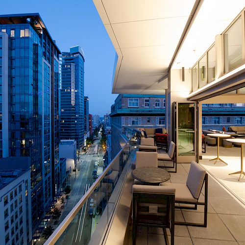 A cityscape view from a modern outdoor balcony at dusk, featuring tall buildings, tables, and seating with warm lighting inside.