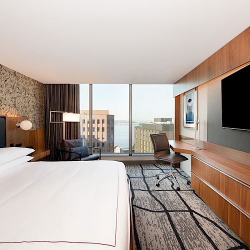 A modern hotel room with a large bed, wall-mounted TV, desk, chair, and window revealing cityscape and water view.