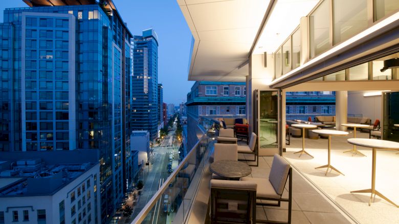 Elevated view of a modern cityscape at dusk, featuring a stylish balcony with seating and open interior space on the right.