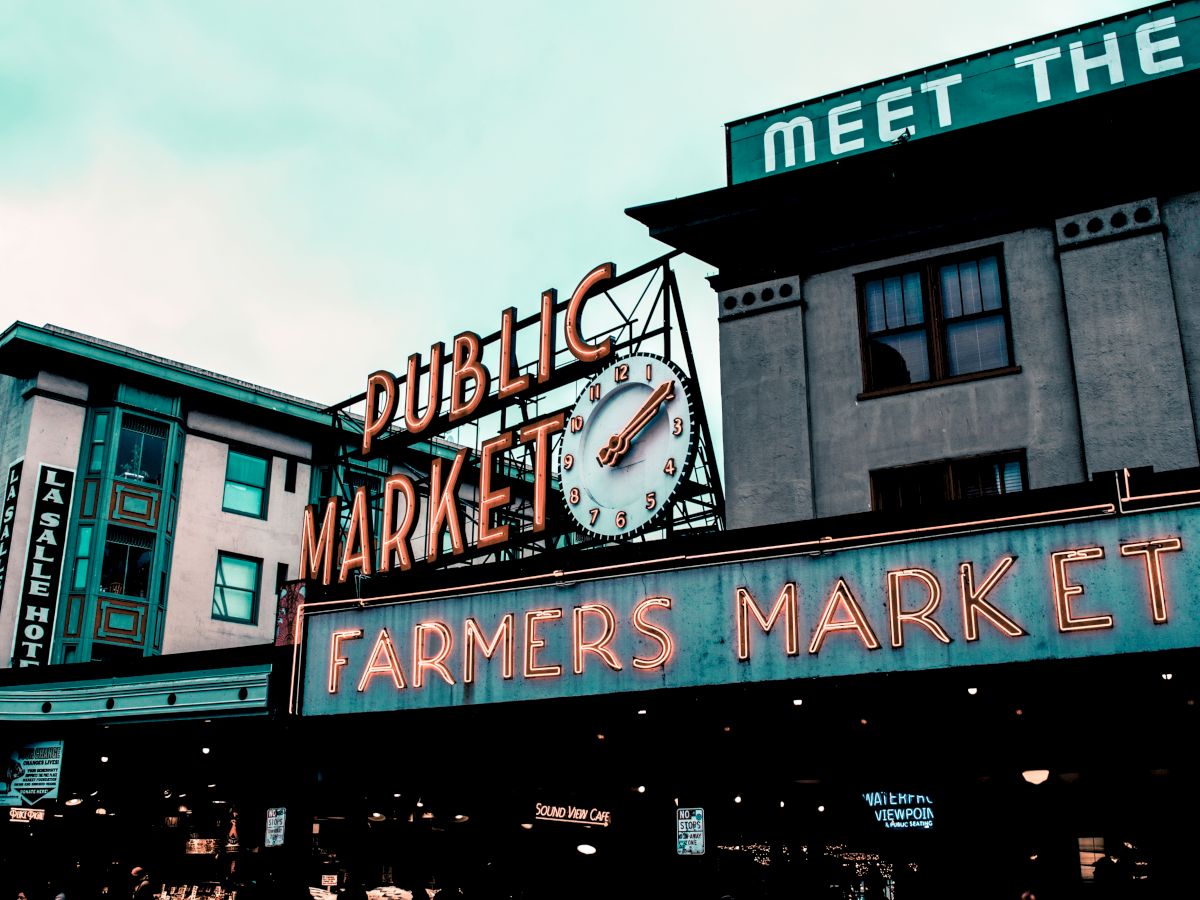 The image shows a neon sign for a "Public Market" and "Farmers Market" with a clock, set against an urban building backdrop.