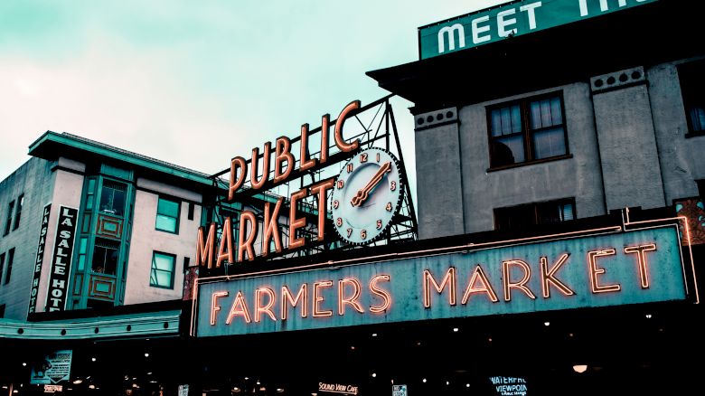 The image shows a neon sign for a "Public Market" and "Farmers Market" with a clock, set against an urban building backdrop.