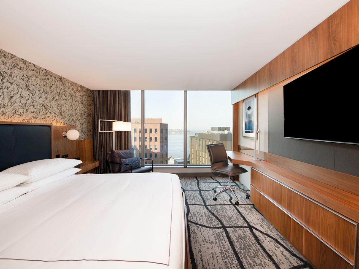 A modern hotel room with a large bed, TV, desk, chair, and a window view of buildings. The decor includes wood panels and patterned wallpaper.