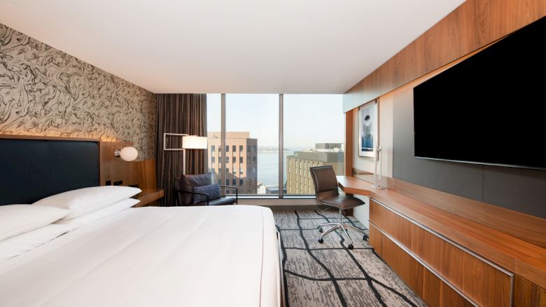 A modern hotel room with a large bed, TV, desk, chair, and a window view of buildings. The decor includes wood panels and patterned wallpaper.