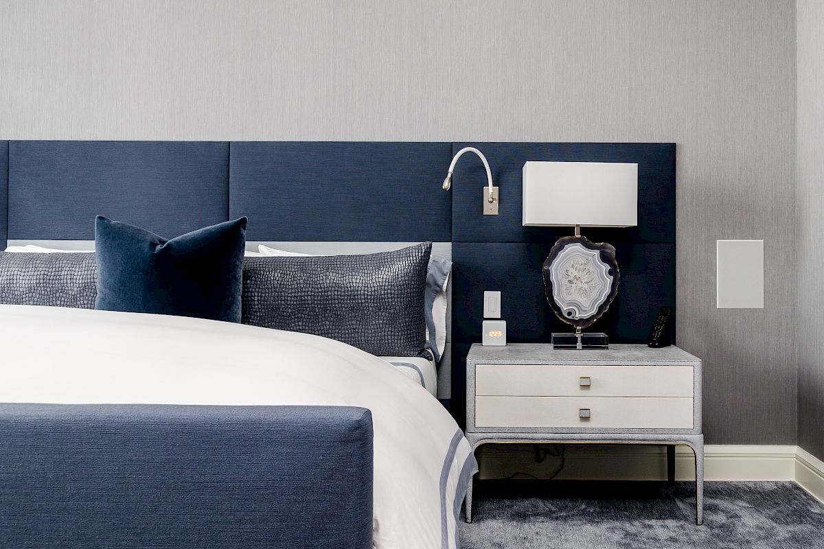A modern bedroom features a navy headboard, white bedding, a side table with a lamp, and a decorative item in a sleek, minimalist design.