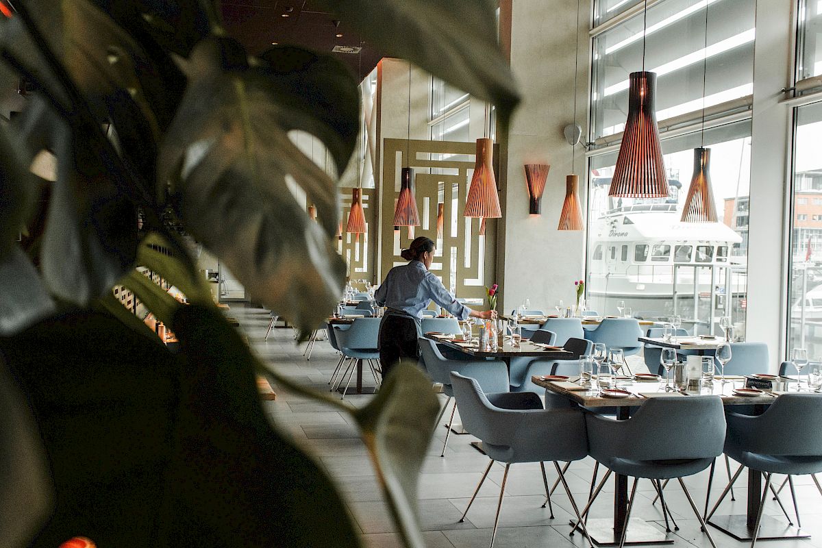 A modern restaurant with sleek design, featuring a server preparing tables and unique pendant lighting. Large windows bring in natural light.