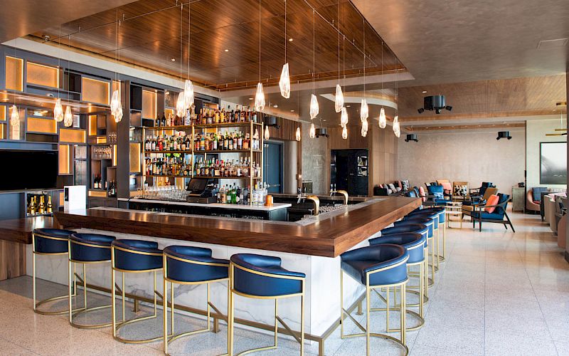 A stylish bar with a wooden counter, blue stools, hanging lights, and a well-stocked liquor shelf is visible. There's a lounge area.