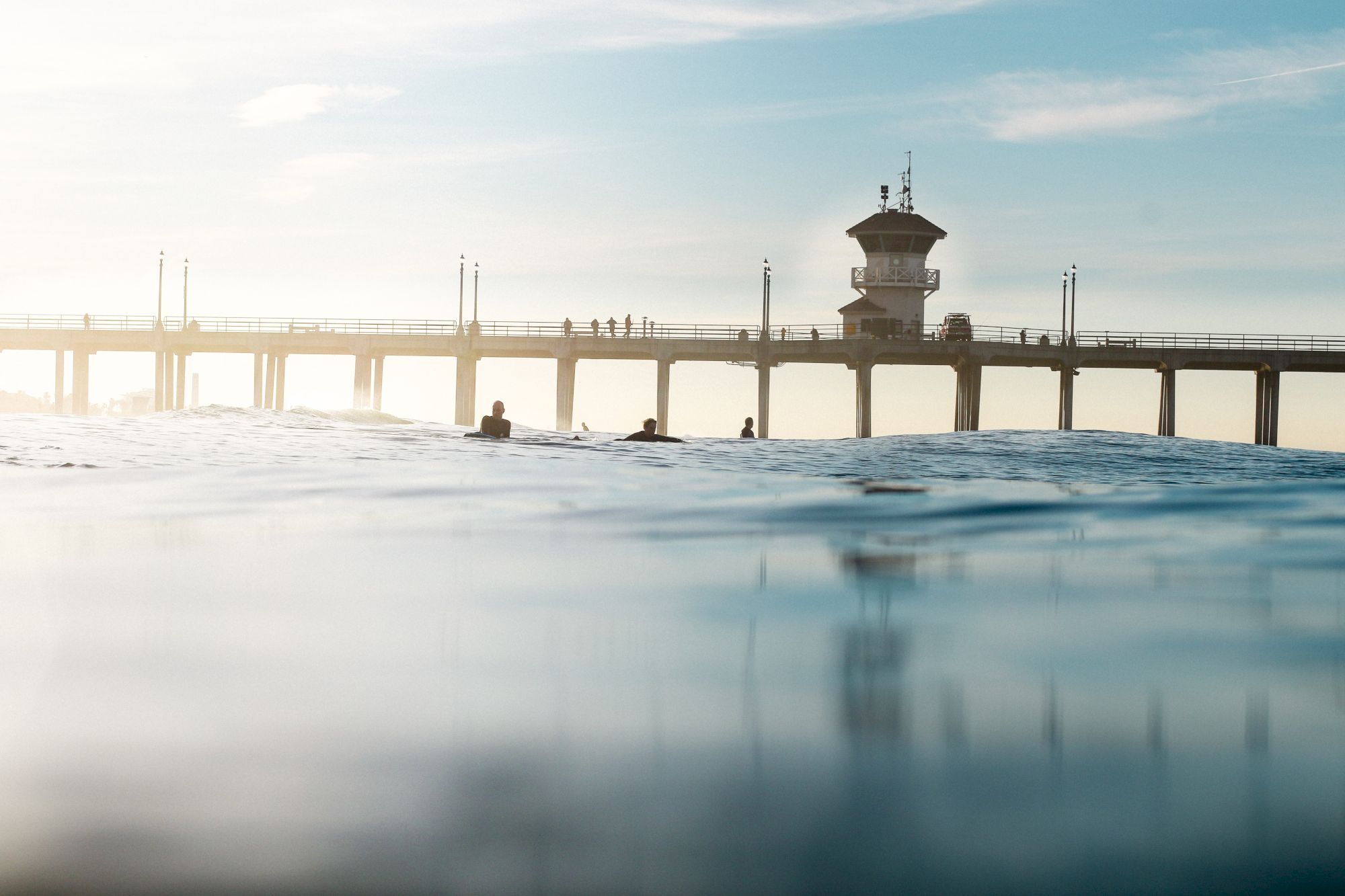 A pier stretches over the water with a few people and a structure visible, and calm water in the foreground under a clear sky.